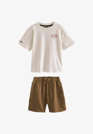 Junge trägt beige T-Shirt, braune Shorts, weiße Turnschuhe, steht und richtet ein kariertes Hemd, das um seine Taille gebunden ist, vor einfachem Hintergrund.