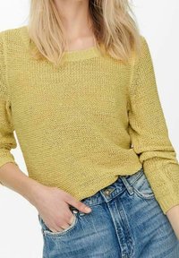 Personne blonde portant un pull jaune texturé avec les manches remontées et un jean bleu taille haute, une main dans la poche.