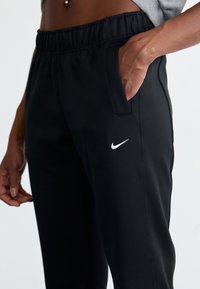 Calças de desporto pretas com um cós elástico, bolsos laterais e um pequeno logótipo branco da Nike na coxa. Textura de tecido suave.