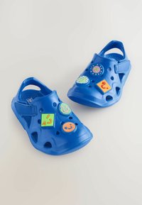 Zoccoli in plastica blu con fori di ventilazione e una fascia in Velcro. Dotati di adesivi decorativi colorati e rimovibili in varie forme.