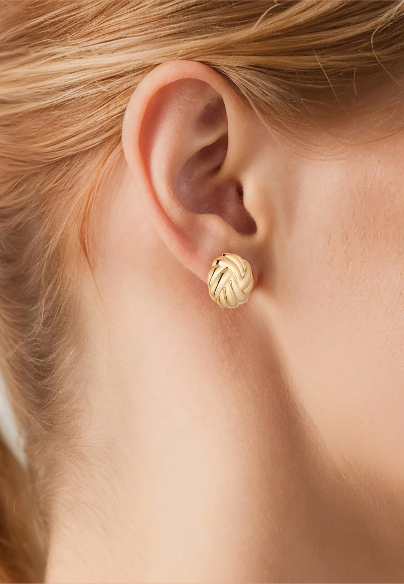 Primer plano de la oreja de una mujer que lleva un pequeño pendiente de botón redondo dorado con un nudo retorcido y el cabello rubio recogido hacia atrás.