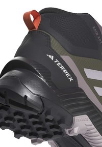 Scarpa da escursionismo nera e oliva con tomaia texturizzata, tallone rinforzato, motivo di battistrada prominente sulla suola e linguetta arancione.