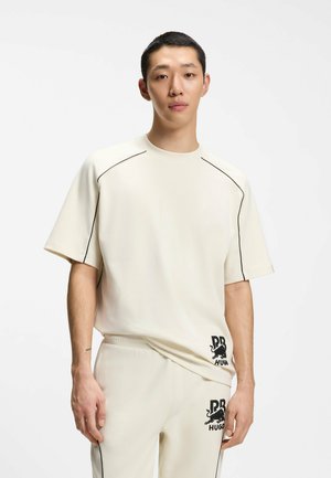 Junger Mann trägt ein off-white kurzärmeliges Hemd und passende Hosen mit schwarzer Paspelierung und "RRHUGO"-Logo auf Hemd und Hose.