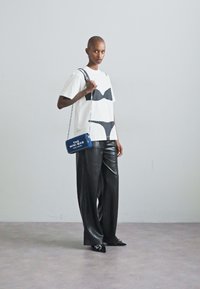 Tricou alb cu modele grafice negre, pantaloni din piele negri, cu croi larg, și o geantă mică albastră tip crossbody cu text alb, purtată împreună cu pantofi negri.