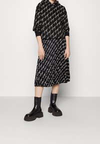 Mujer vistiendo una sudadera negra con capucha y una falda midi plisada con un guion repetido en blanco, combinada con botas de plataforma gruesas negras.