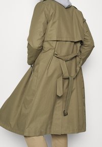 Persona vistiendo un abrigo trinchero de color verde oliva con un cinturón atado en la parte trasera, pantalones beige y una sudadera gris claro de capa debajo.