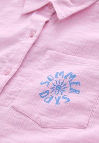 Chemise boutonnée rose clair en tissu texturé, avec une poche sur la poitrine gauche et un graphique bleu "SUMMER DAYS" avec un détail soleil.