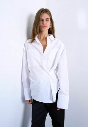 Femme aux cheveux mi-longs portant une chemise blanche oversize boutonnée et un pantalon noir, debout devant un mur clair uni.