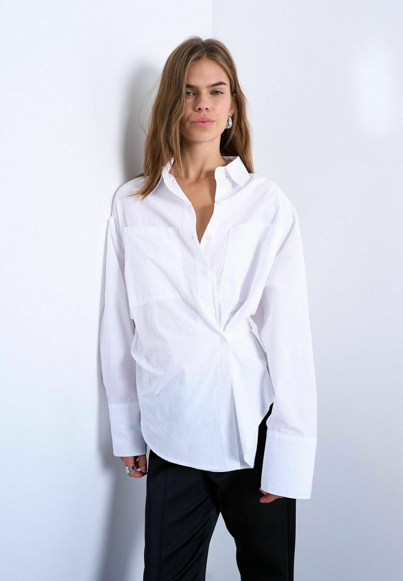 Femme aux cheveux mi-longs portant une chemise blanche oversize boutonnée et un pantalon noir, debout devant un mur clair uni.