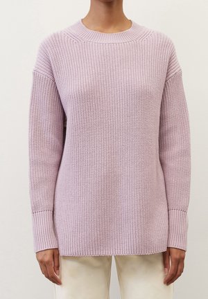 Kobieta w jasnofioletowym prążkowanym swetrze z długimi rękawami i okrągłym dekoltem, w połączeniu z jasnobeżowymi spodniami.