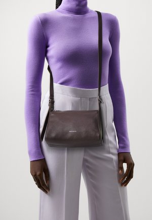 Brun läder crossbody-väskan med en texturerad yta, har en dragkedjestängning och justerbar rem. Bärs över en lila topp och ljusa byxor.