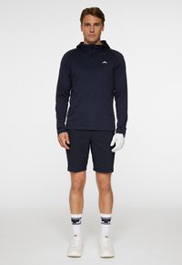 Marinblå långärmad tröja med huvtröja, dragkedja vid kragen och logotyp; kombinerad med marinblå shorts, vita handskar och vita sneakers, samt randiga strumpor.