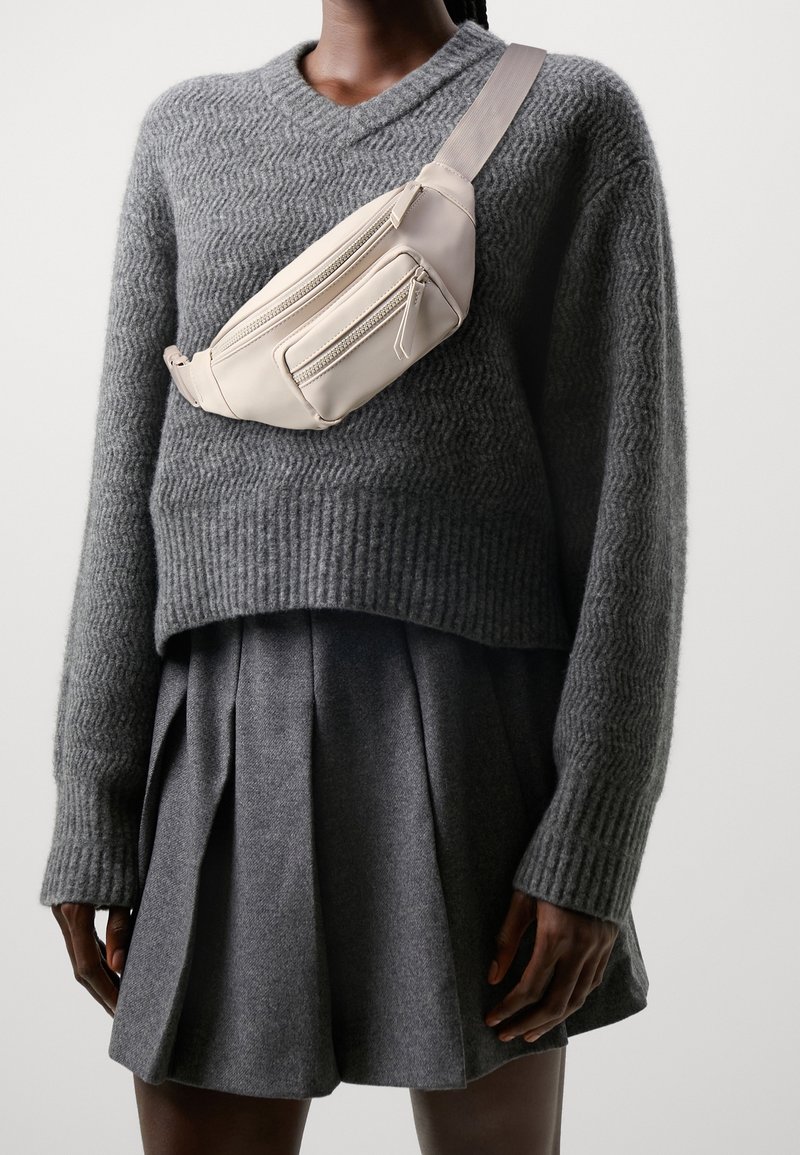 Béžová nylonová ledvinka se dvěma kapsami na zip, nošená přes rameno přes texturovaný šedý svetr a šedou plisovanou sukni na míru.