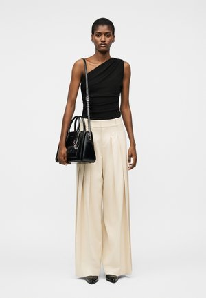 Žena v čiernom topu na jedno rameno, béžové široké plisované nohavice a čierne špicaté topánky, drží čiernu kabelku so zaveseným pásikom na ramene.