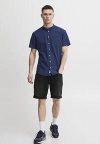 Cămașă cu mânecă scurtă, de culoare bleu marin, cu nasturi, dintr-un material ușor, având un buzunar în față, asortată cu pantaloni scurți negri răsuciți și pantofi sport.