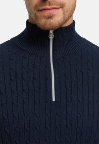 Marineblå kabelstrikket genser med glidelås og høy krage, med en sølvfarget glidelås og strukturert strikkemønster. Elastisk, varm stoff som passer godt for lag-på-lag