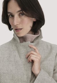 Hellgrauer Wollmantel mit hohem Kragen und feiner Textur. Das Modell trägt darunter einen rosa gerippten Rollkragenpullover.