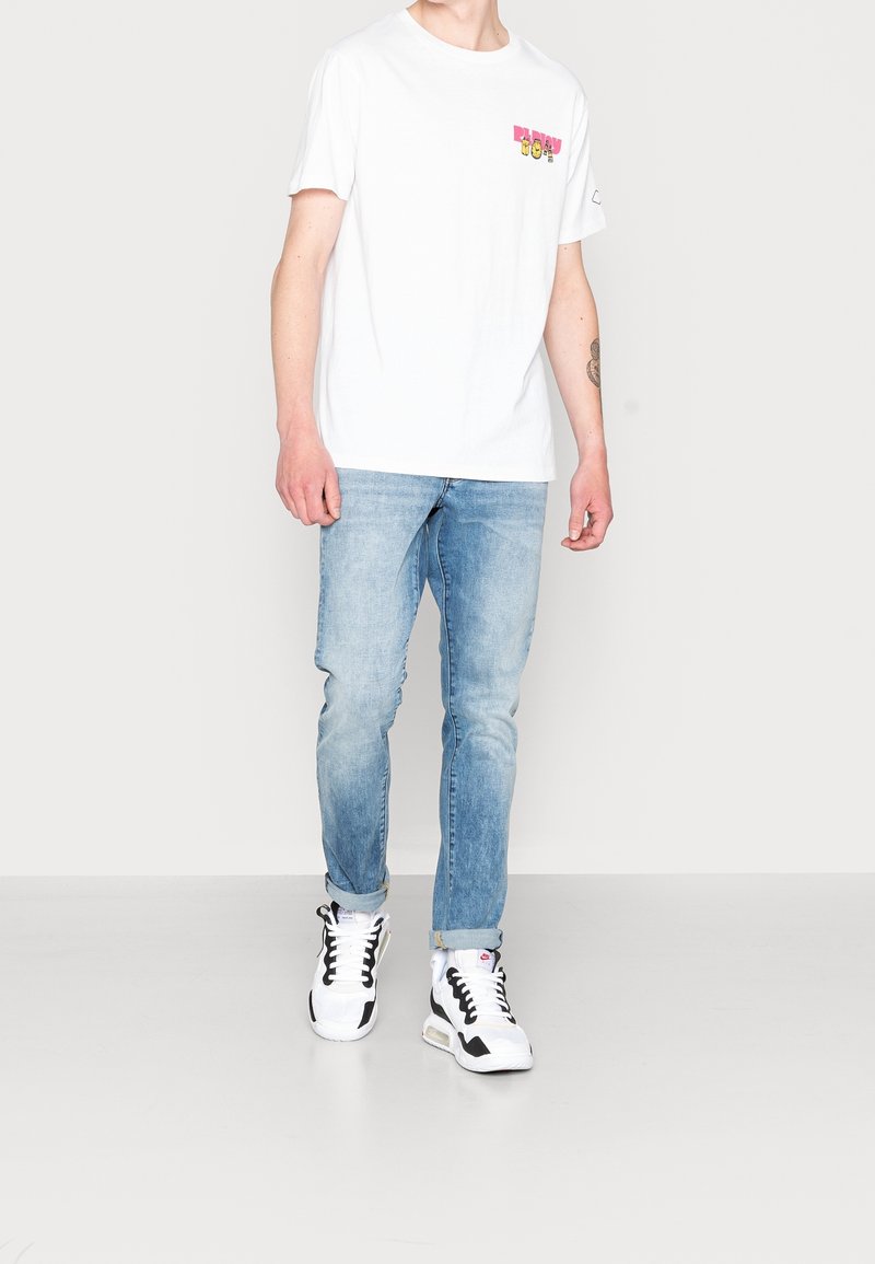 Hvid t-shirt med korte ærmer og farverigt frontmotiv, parret med lyseblå tætsiddende jeans og sort-hvide sportsko.