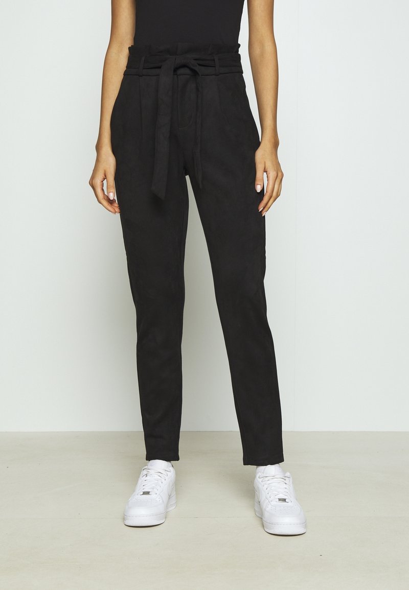 Femme portant un pantalon noir taille haute noué et des baskets blanches, debout contre un fond clair uni.