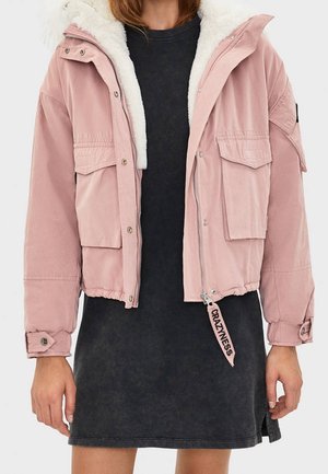 Pink, cropped bomber jacket with fur-lined hood, zip closure, multiple front pockets, and elastic cuffs. Worn over a dark dress.