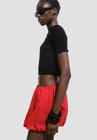 Top scurt negru cu dungi, pantaloni scurți roșii tip balon și brățări negre suprapuse. Model purtând ochelari de soare negri supradimensionați; profil lateral pe un fundal simplu.