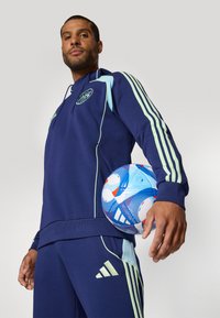 Navy treningsdrakt med lyseblå detaljer og tre striper, med en logo på brystet; modellen holder en flerfarget fotball.