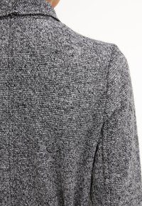 Gros plan sur la zone de l'épaule arrière d'une personne portant une veste tricotée grise texturée avec une couture et un col visibles.