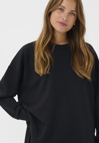 Vrouw met lang lichtbruin haar, gekleed in een losse zwarte sweater, staande met één hand op haar heup tegen een effen lichte achtergrond.