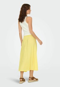 Top blanco de tirantes combinado con una larga falda amarilla de lino. La falda tiene una cintura elástica y una textura suave. Sandalias marrones con tacón de cuña.