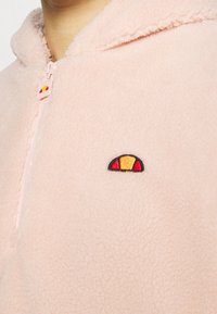 Vêtement en polaire rose clair avec une fermeture éclair à moitié et un petit logo rond jaune et rouge sur la poitrine.