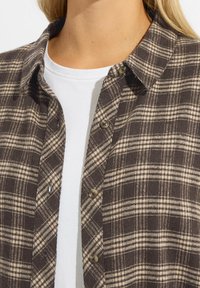 Camicia di flanella a quadri marrone e beige con button-down e una maglietta bianca a girocollo sotto. Tessuto morbido, design informale.