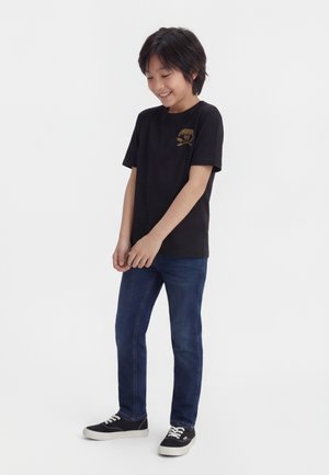 Garçon souriant portant un t-shirt graphique noir à manches courtes, un jean bleu et des baskets noires, debout les mains jointes devant lui.