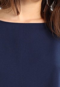Haut bleu marine à épaule dénudée en tissu lisse, avec un col épuré et sans motifs ni accessoires distincts.