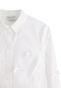 Camicia bianca in cotone con colletto a punta, decorata con fiocchi bianchi testurizzati e chiusura con bottoni. Design semplice e pulito con una finitura morbida.