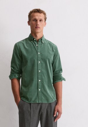 Man with curly hair wearing green button-up shirt with rolled sleeves and gray pants, standing against a white textured wall.