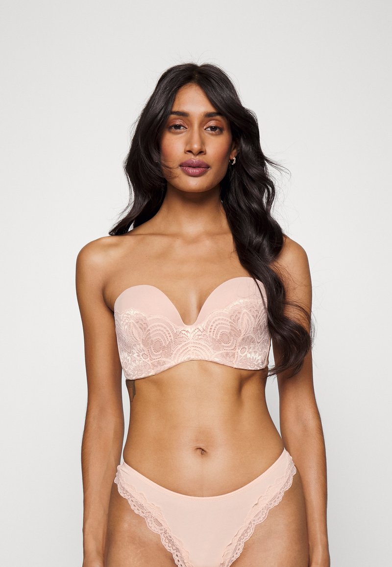 Bralette uten stropper i myk rosa med blonderdetaljer, glatte kopper og dyp V-form. Matchende truser med blondekanter kompletterer settet.