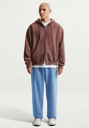 Hombre con sudadera marrón con cremallera, camiseta blanca, pantalones deportivos azul claro y zapatillas blancas, de pie frente a un fondo liso.