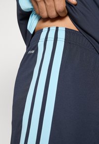 Calções de atletismo azuis marinhos com três riscas verticais azul claro de cada lado, feitos de um tecido leve com uma textura suave.