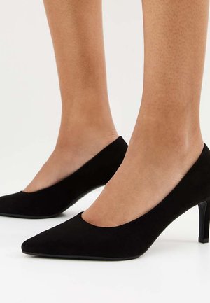 Voeten met zwarte puntschoenen met lage kittenhakken op een witte vloer, met zijaanzichten van beide schoenen en enkels.