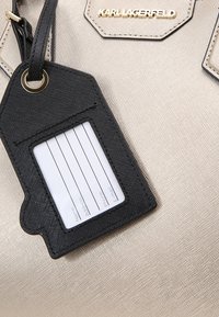 Beige handväska med en strukturerad yta, med en svart läderbagagetagg som har ett vitt identitetskort och guldlogotypdetalj.