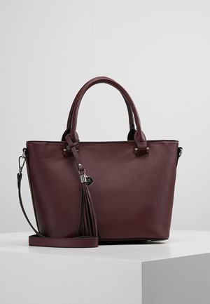 Dunkelburgunderfarbene strukturierte Tragetasche mit zwei Griffen, abnehmbarem Schulterriemen und dekorativem Quastenanhänger auf einem weißen Podest.