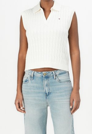 Personne portant un pull polo sans manches en maille torsadée blanche et un jean taille haute bleu clair sur un fond blanc uni.