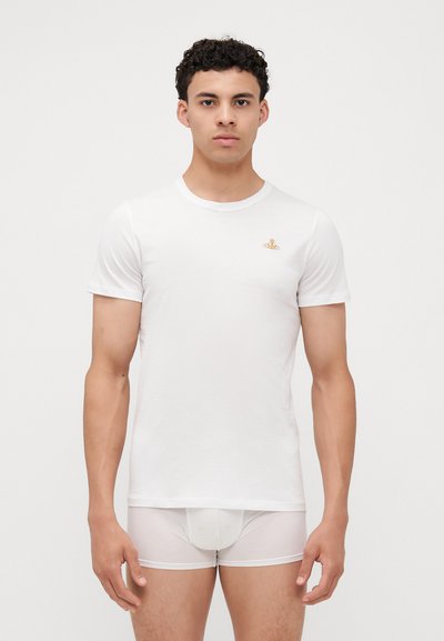 Camiseta blanca de algodón de manga corta con cuello redondo, que presenta un pequeño logotipo bordado en oro. Combinada con unas braguitas blancas transparentes.