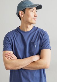 T-shirt de algodão azul-marinho com um bolso no lado esquerdo do peito e logótipo bordado. Acompanhado por um boné de textura azul claro com aba curva.