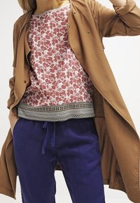 Lichte bruine jas over een top met bloemmotief in rood en wit, met een zwart-wit gestreepte zoom; gecombineerd met blauwe broek met aantrekkoord.