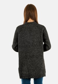 Kvinde med langt brunt h�år iført en mørkegrå striktrøje og blå jeans, vist fra ryggen mod en hvid baggrund.