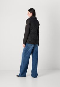 Vrouw met lang haar draagt een zwarte gewatteerde vest en losse blauwe jeans, staande met haar rug naar de camera tegen een effen achtergrond.