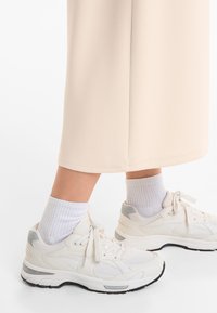 White sneakers with mesh and synthetic materials, featuring gray accents and a textured sole, paired with ribbed white socks and a beige garment.