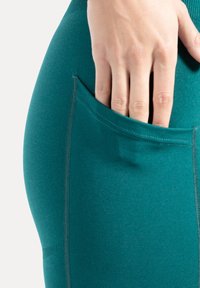 Leggings turquoise en tissu extensible, avec une poche latérale et des détails de couture apparents. Texture lisse avec une coupe ajustée.