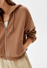 Kvinna som bär en kort brun zip-up hoodie med elastiska ärmslut och nederkant, handen i fickan, matchad med beiga byxor mot en neutral bakgrund.